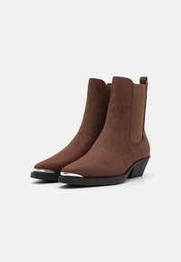 Braune Wildleder-Stiefeletten mit elastischen Seitenteilen, spitzer Zehenform und metallischen Akzenten auf der Zehenkappe. Niedriger Blockabsatz.