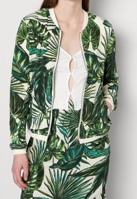 Vrouw in een tropische bladerprint jacket en rok met een wit cropped knooptop, handen in de zakken van het jacket, tegen een eenvoudige achtergrond.