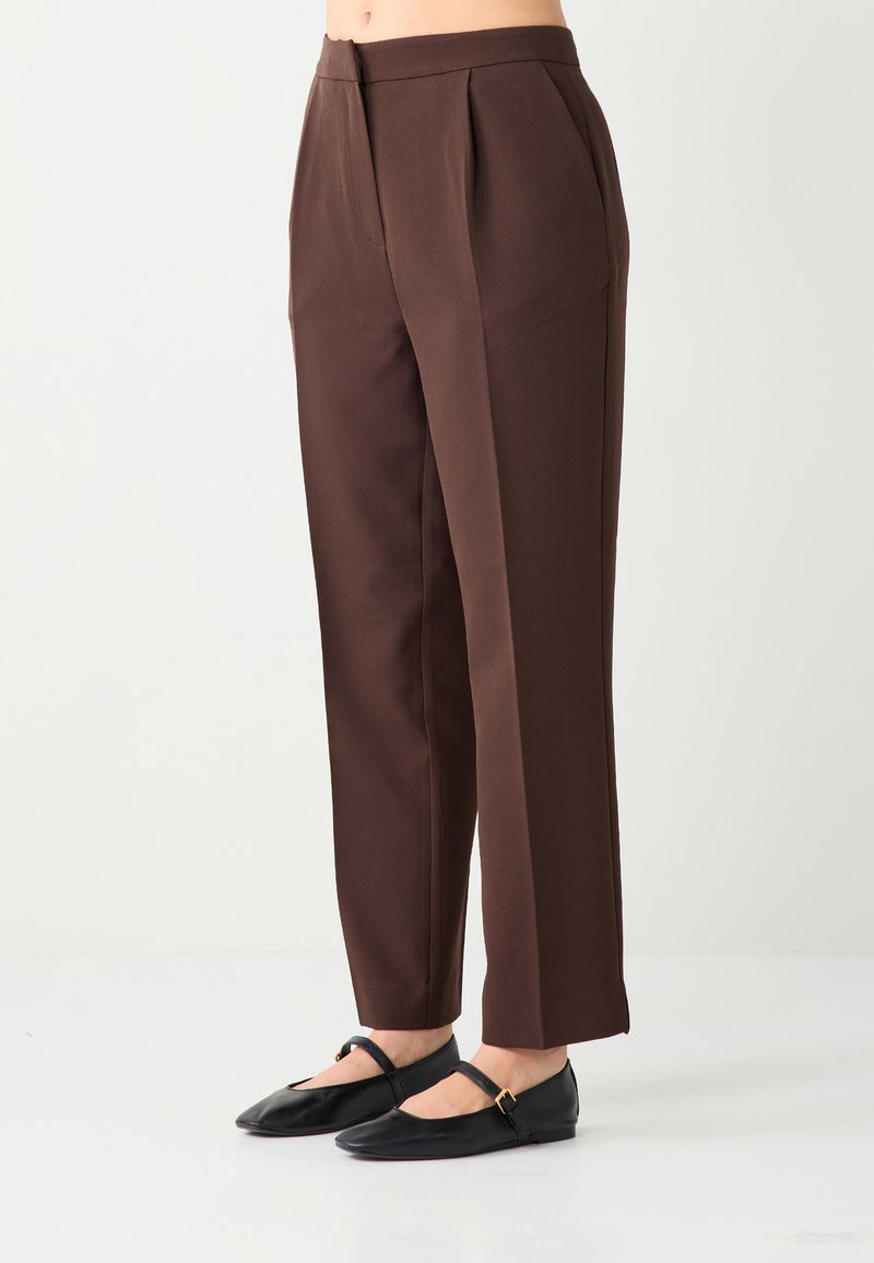 Pantalon marron sur mesure avec plis, coupe droite et texture lisse. Associé à des ballerines Mary Jane noires, présentant un détail boucle.