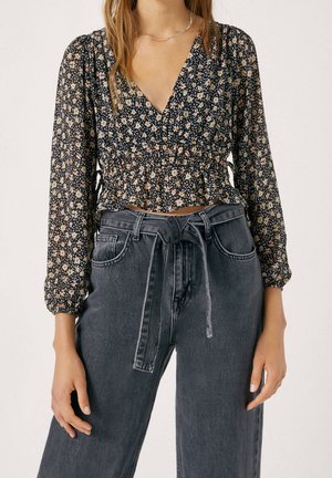 Femme portant un chemisier noir à manches longues à motif floral et col en V, rentrée dans un jean noir taille haute avec une ceinture nouée à la taille.
