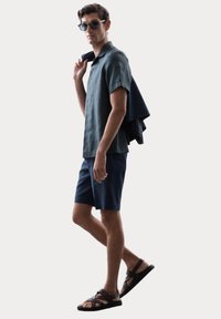 Herrenoutfit mit einem dunkelgrünen kurzärmligen Leinenhemd, marineblauen Shorts und braunen Sandalen mit Riemen. Accessoire: dunkle Sonnenbrille.
