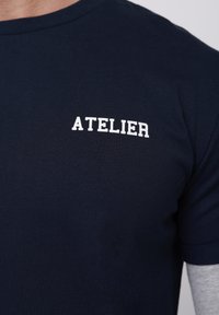 Tumši zila īsroku kokvilnas T-krekls ar vārdu "ATELIER" baltos burtiem kreisajā krūtīs.