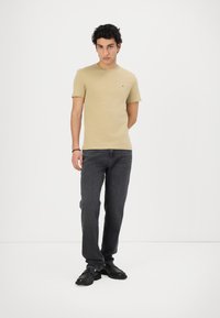 T-shirt beige in cotone con colletto rotondo e piccolo logo sul petto, abbinata a jeans grigio scuro e scarpe nere con suola spessa.