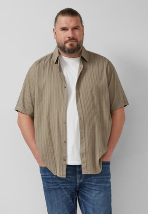 Bärtiger Mann mit zurückgebundenen Haaren, trägt ein offenes beiges Kurzarmhemd über einem weißen T-Shirt und blaue Jeans, steht mit den Händen in den Taschen.