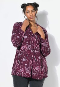 Vrouw met donker haar in twee knotten, gekleed in een paarse paisley knoopshirt en donkere broek, poseert tegen een witte achtergrond.