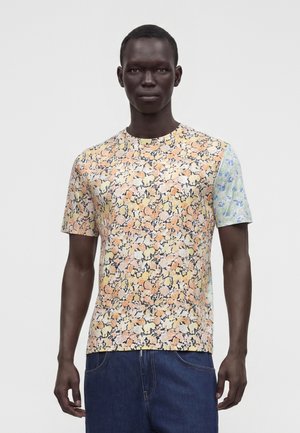Bărbat purtând un tricou cu model floral multicolor cu mâneci scurte și blugi albaștri, stând în picioare în fața unui fundal simplu și deschis la culoare.
