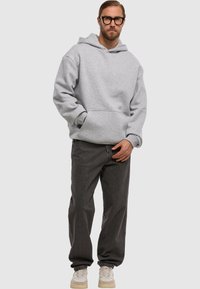 Man in zwartomrande bril, lichtgrijze hoodie, donkergrijze relaxte jeans en witte sneakers, staand met één hand in zijn zak.