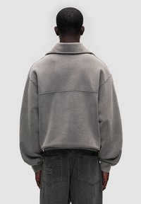 Šedá oversized mikina s polo límečkem, s prošívanými detaily na zadní straně, doplněná tmavě šedými džíny s vyšitým logem.