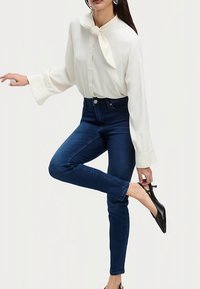 Bílá halenka s límečkem vázaným na mašli, předními knoflíky a rozšířenými rukávy, kombinovaná s tmavě modrými džíny s vysokým pasem a černými podpatky.