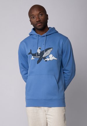 Blauwe hoodie van zacht materiaal, met een afbeelding van een astronaut die op een walvis rijdt, omgeven door wolken. Voorzien van een kangoeroezak en een handschoen met trekkoord.