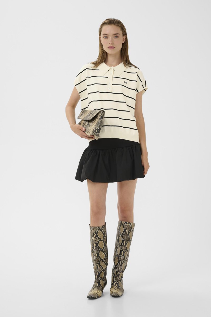 Kvinde iført hvidstribet polo shirt, sort mininederdel, holder en clutch af slangeskind, iført knæhøje støvler af slangeskind, stående på ensfarvet baggrund.