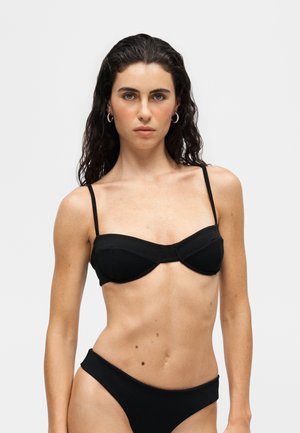 Młoda kobieta z długimi ciemnymi kręconymi włosami, ubrana w czarny top i dół od bikini na jasnym jednolitym tle.