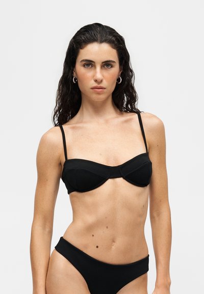 Jeune femme aux longs cheveux bouclés et foncés portant un haut et un bas de bikini noirs sur un fond clair uni.