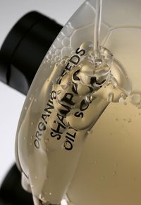Botella de champú transparente llena de un líquido amarillo claro, con burbujas y texto negro que la etiqueta como "Champú Orgánico de Semillas para Cuero Cabelludo Graso".