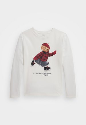 Camisola de manga longa branca com uma gráfica de um urso vestido com uma camisa de quadros vermelha, saia cinza e sapatos pretos, com assinatura impressa abaixo.