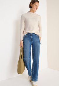 Jasno beżowy sweter dzianinowy z wysokim kołnierzem, zestawiony z niebieskimi spodniami typu straight-leg i zieloną torbą na ramię. Białe trampki z detalami tekstury.