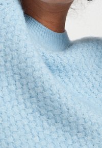 Pull en maille texturée bleu clair avec un col rond côtelé, un tissu doux et une coupe ample, mettant en valeur des motifs de tissage complexes.
