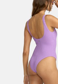 Maillot de bain une pièce violet avec un tissu texturé, dos rond et larges bretelles. Présente un design à jambe haute et un détail de branding minimal.
