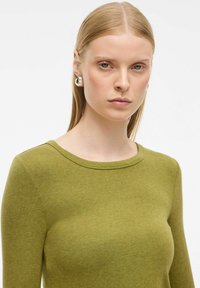 Zeleni dolg rokav s okroglim izrezom, gladko teksturo in prilegajočim dizajnom, v kombinaciji z velikimi kovinskimi okroglimi uhani.