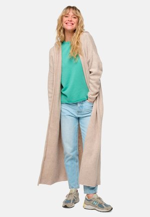 Long beige cardigan, soft texture, open front, layered over green t-shirt, light blue jeans, and gray sneakers with blue accents.