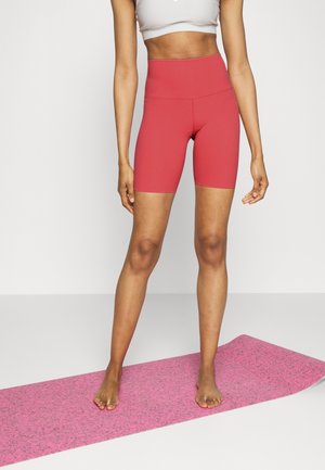 Frau in korallenroten, hoch taillierten Biker-Shorts und weißem bauchfreiem Top, steht barfuß auf einer pinkfarbenen Yogamatte vor einem einfarbigen Hintergrund.