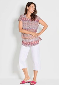 Kurze Ärmel, gemusterte Bluse in Rosa, Lila und Blau, kombiniert mit weißen, taillierten Hosen und rosa geflochtenen Sandalen.