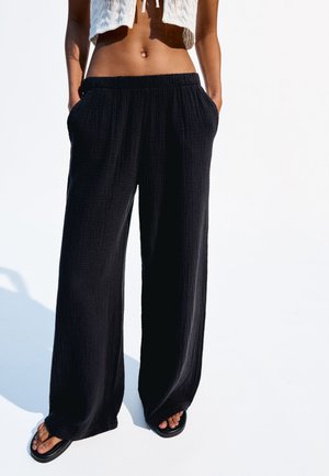 Pantalons noirs à jambes larges en tissu texturé, dotés d'une taille élastique et de poches latérales, associés à des tongs noires simples.