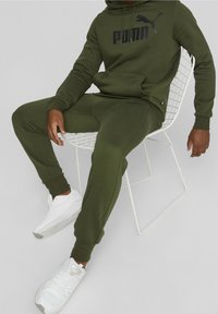 Olivgrön huvtröja och joggerset i mjukt material, med en svart logotyp på framsidan. Bärs med vita sneakers, sittande på en trådstol.