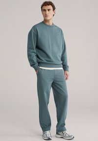 Tirkizno zelena dukserica i odgovarajuće trenirke od mekanog materijala, s jednostavnim dizajnom i rebrastim manžetnama, u paru s laganim tenisicama.