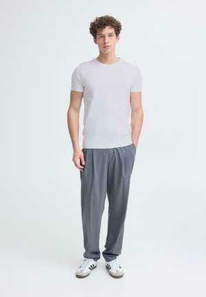 Jonge man met krullend haar, gekleed in een effen wit T-shirt, grijze geplooide broek en witte sneakers, staand tegen een lichte achtergrond.