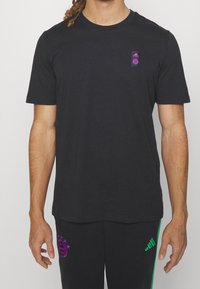 Schwarzes T-Shirt mit kurzen Ärmeln aus Baumwolle und rundem Ausschnitt, mit einem lila Adidas-Logo auf der Brust. Kombiniert mit schwarzen Hosen.