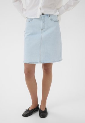 Frau trägt einen hellblauen Jeansrock in Knielänge, kombiniert mit schwarzen Ballerinas und einem weißen Langarmhemd.