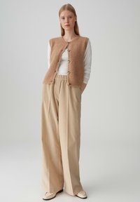 Gilet lavorato a maglia beige con bottoni sopra una camicia bianca a maniche lunghe, abbinato a pantaloni di corduroy beige a vita alta e larghi. Texture liscia.