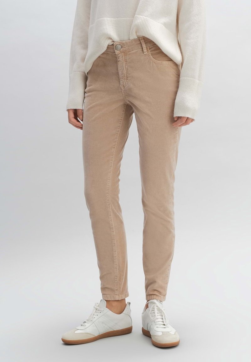 Pantalon en velours côtelé beige à coupe slim, avec une fermeture à boutons sur le devant et un petit logo brodé, porté avec des baskets blanches.