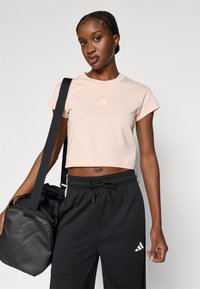 Ροζ ανοιχτόχρωμη κοντή μπλούζα με κοντά μανίκια και μικρό λογότυπο στο στήθος, σε συνδυασμό με μαύρο αθλητικό παντελόνι από δίχτυ και μαύρη τσάντα duffel.