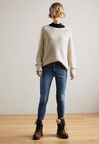 Crème ribgebreide trui, zwarte binnenlaag, blauwe slim jeans en bruine laarzen met bontaccenten, staand op een houten vloer.