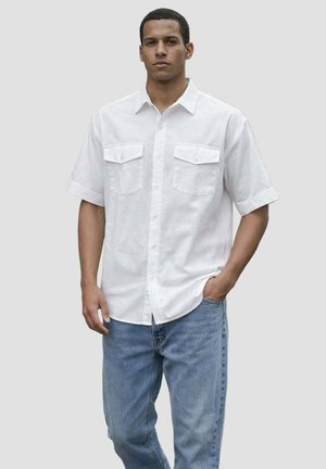 Homme portant une chemise blanche à manches courtes avec deux poches poitrine et un jean bleu clair, debout avec une main dans la poche.