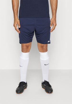 Marineblå atletiske shorts med hvidt "JAK"-logo, kombineret med hvide knæstrømper med sort "Nike" swoosh og sorte fodboldstøvler.