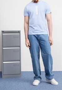 Světle modré tričko s logem "ALPHA", doplněné volnými modrými džíny s cargo kapsami. Bílé tenisky a šedý kovový spisdní skříň v pozadí.