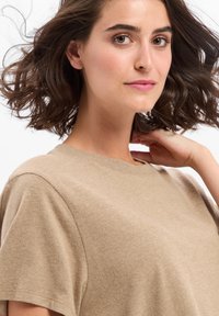 Beige katoenen t-shirt met een ronde halslijn en korte mouwen, met een zachte textuur en een relaxte pasvorm. Het haar is in losse golven gestyled.