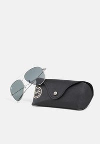 Ochelari de soare aviator metalici cu lentile gri, așezati lângă o carcasă din piele neagră, care prezintă un logo circular și un sistem de închidere tip snap.