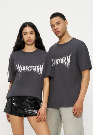 Δυο νεαροί ενήλικες στέκονται δίπλα-δίπλα φορώντας σκούρα γκρι μπλουζάκια με το λογότυπο "Yourturn"· ο ένας συνδυάζει τη μπλούζα με μαύρα σορτς, ο άλλος με ανοιχτόχρωμο τζιν.