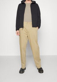 Svart zip-up hoodie, beige t-shirt, beige lösa byxor och svarta skor. Tyget har en mjuk textur och en minimalt designad struktur.