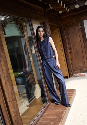 Dvoudílný outfit v tmavém denimovém provedení. Zkrácený top bez rukávů s kulatým výstřihem a detailem s knoflíkem, spárovaný s volnými kalhotami s vysokým pasem.