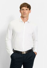 Weißes Langarmhemd mit Button-Down-Kragen, kombiniert mit dunkler Hose und einem schwarzen Gürtel mit rechteckiger silberner Schnalle.