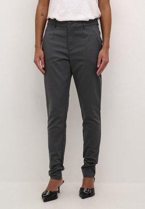 Frau trägt dunkelgraue, maßgeschneiderte Hose, weißes Oberteil und schwarze spitze High Heels mit dekorativen Schnallen, steht vor einem schlichten Hintergrund.