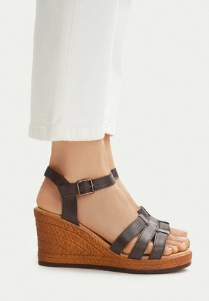 Persona que lleva sandalias cuña de cuero marrón con correas de hebilla en el tobillo y tacones de corcho texturizados, combinadas con pantalones blancos recortados.