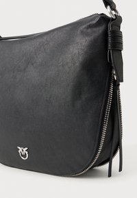 Melna ādas pleca soma ar sudraba rāvējslēdzēju un mazu sudraba putna logotipu priekšpusē, uz baltā fona.