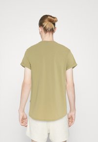 Olivově zelené tričko vyrobené z měkké bavlny, s krátkými ohrnutými rukávy a zaobleným lemem. Pohled zezadu ukazuje hladkou texturu a minimální design.