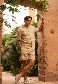 Lichtbeige set van een kortarmige shirt en shorts met een wit bladmotief. Draag met grijze instappers. Achtergrond toont bomen en gestructureerde muren.